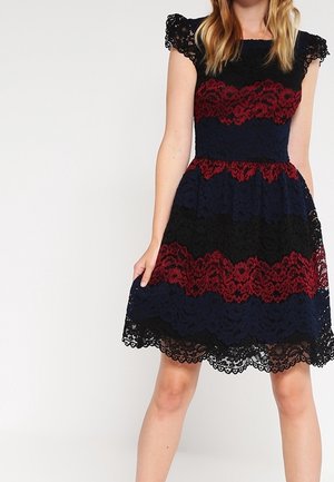 Kanten jurk met een aangesloten lijfje, korte kapmouwen en een uitlopende rok. In kleurvlakken van marineblauw, rood en zwart met een bloemenpatroon.