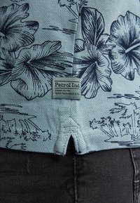 Modrý textil s květinovým vzorem a námořnickými ilustracemi, na dolním levém rohu s nálepkou "Petrol Ind" a s výřezovým designem na lemu.