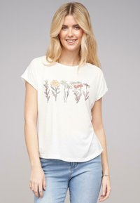 T-shirt crème avec des manches courtes retroussées, ornée d'un motif floral dans des couleurs douces sur la poitrine. Coupe douce et décontractée.