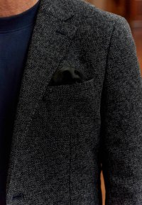 Blazer gris texturizado hecho de tejido tejido, con solapa de muesca y un pañuelo negro en el bolsillo. Camisa azul de cuello redondo debajo.