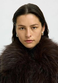 Abrigo de piel oscura con un cuello voluminoso, llevado sobre un suéter negro. El modelo tiene el cabello oscuro de longitud media y aretes de aro plateados.