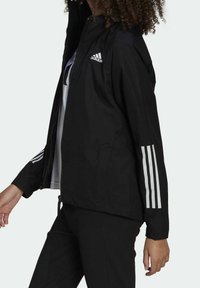 Giacca leggera nera con zip frontale, cappuccio e tre strisce bianche sulle maniche. Presenta un logo Adidas sul petto.