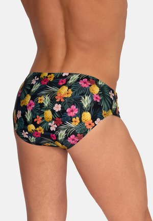 Uomo che indossa slip da bagno neri con motivo colorato tropicale di fiori e ananas, mostrato dalla metà della schiena fino a metà coscia su sfondo semplice.