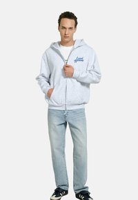 Young man wearing a light gray zip-up hoodie with "Local Heroes" text, light blue jeans, and black sneakers, standing with one hand in pocket.