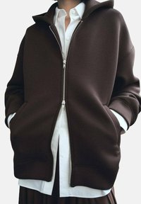 Sudadera marrón con cremallera hecha de una tela suave, con un cuello alto, grandes bolsillos laterales y puños de canalé, superpuesta sobre una camisa blanca.
