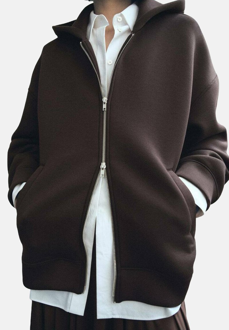 Sudadera marrón con cremallera hecha de una tela suave, con un cuello alto, grandes bolsillos laterales y puños de canalé, superpuesta sobre una camisa blanca.