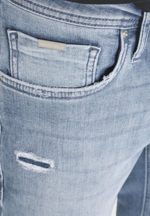 Džep od svijetloplavih traperica s malom metalnom naljepnicom marke i naboranim horizontalnim oštećenjem ispod ruba džepa.