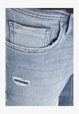 Džep od svijetloplavih traperica s malom metalnom naljepnicom marke i naboranim horizontalnim oštećenjem ispod ruba džepa.