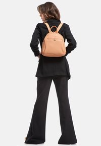 Zaino in pelle color pesco chiaro con finitura testurizzata, spallacci doppi, manico superiore e tasca anteriore, indossato da un modello che indossa un outfit nero.