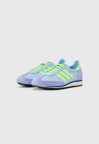 Zapatillas deportivas con una mezcla de ante azul claro y púrpura, con cordones de neón verde y tres franjas en el lateral. Suela de goma con tracción.