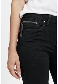 Acercamiento a una persona que lleva puestos jeans negros con un bolsillo delantero con cremallera y una camisa blanca metida por dentro, mostrando la cadera izquierda y la mano.