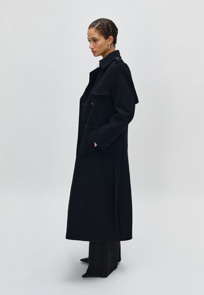 Lungo cappotto in lana nera con ampio colletto, tasche laterali e dettagli botones. Il modello indossa pantaloni neri e stivali a punta. Design semplice ed elegante.