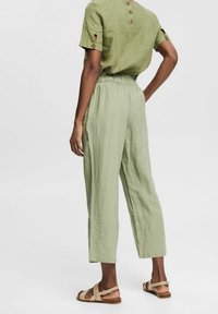 Persona con pantalones de lino verde claro con cintura elástica, blusa de manga corta a juego con detalles de botones y sandalias planas beige, de pie de lado.