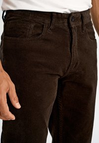 Brązowe spodnie sztruksowe z teksturowanym, prążkowanym wzorem, pięcioma kieszeniami i zapięciem na guzik. Ręka delikatnie dotyka materiału.