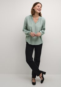 Blusa de manga larga verde claro con escote en V y puños elásticos, con un sutil estampado a cuadros. Combinada con pantalones negros de pierna recta y tacones.