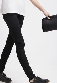 Černé úzké džíny vyrobené z elastického materiálu se hladkým povrchem, doplněné černými šněrovacími botami a černou koženou kabelkou.