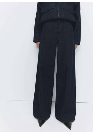 Personne portant un pantalon noir ample à jambes larges et des chaussures pointues noires, debout sur un fond blanc.