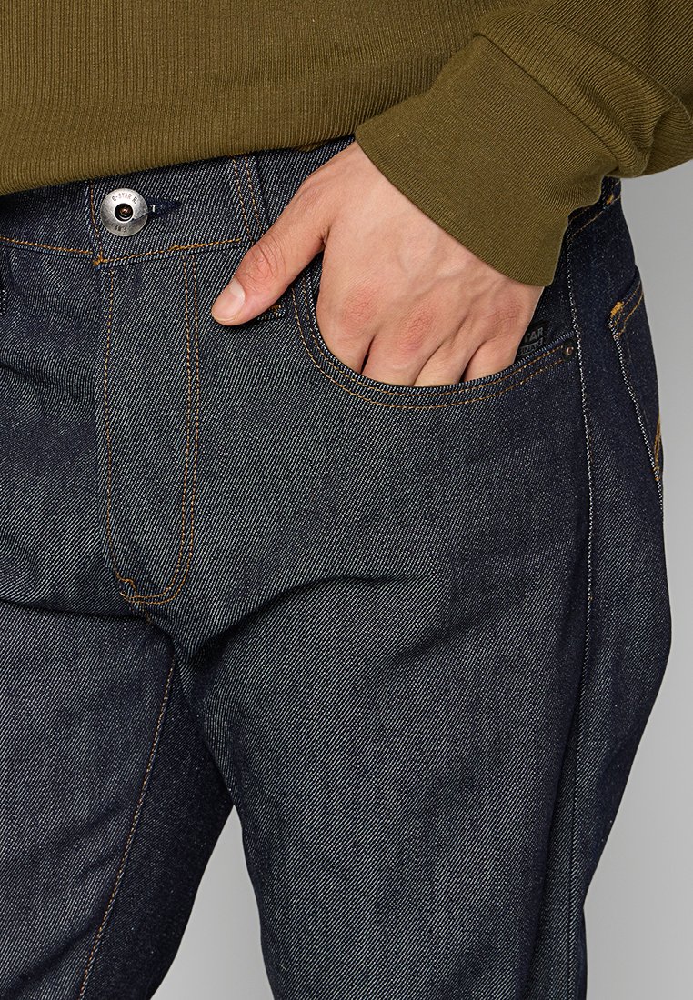 Dunkelblaue Jeans mit orangefarbener Naht, einem silbernen Knopfverschluss und einer Seitentasche, präsentiert mit einer Hand, die in der Tasche ruht.