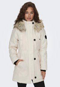 Lichtbeige parka met een nepbont gevoerde capuchon, zwarte knopen, een elastische taille en twee zijzakken. Zachte textuur en een middellange ontwerp.