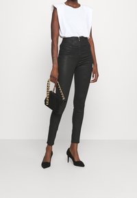 Mujer vistiendo una blusa sin mangas blanca, pantalones ajustados negros de cintura alta, tacones altos negros, sosteniendo un bolso negro con cadenas doradas.