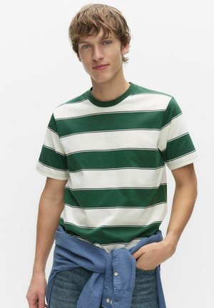 Ung man med grön- och vitrandig t-shirt och en blå skjorta knuten runt midjan, stående mot en enfärgad vit bakgrund.