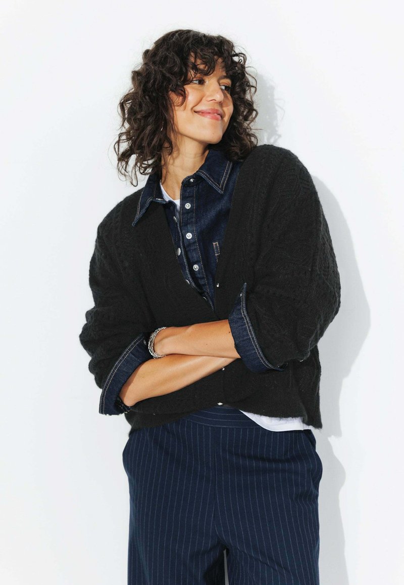 Cardigan noir texturé sur une chemise en denim avec un t-shirt blanc en dessous, associé à un pantalon bleu marine à rayures. Le mannequin pose les bras croisés.