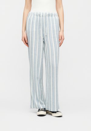 Person som har på seg løse, høytlivede bukser med vertikale blå og hvite striper og svart-hvite joggesko, står vendt mot fronten.