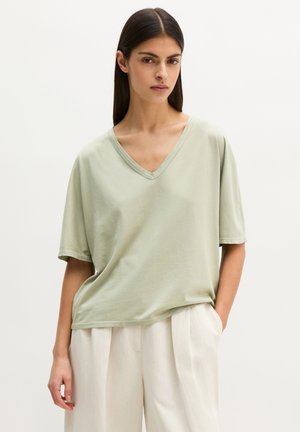 Kobieta o długich ciemnych włosach, ubrana w luźny jasnooliwkowy T-shirt z dekoltem w serek oraz białe, plisowane spodnie z wysokim stanem, z ręką w kieszeni.