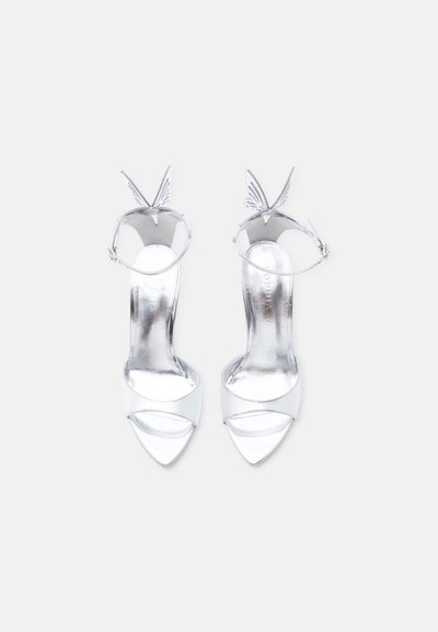 Ζευγάρι λαμπερών ασημένιων σανδαλιών με ψηλά τακούνια, λουράκια στον αστράγαλο και λεπτομέρειες σε σχήμα φτερού, σε λευκό φόντο.