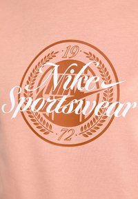 Lys rosa bomull T-skjorte med et rundt brunt emblem som har teksten "Nike Sportswear" og tallene "19" og "72" inni.