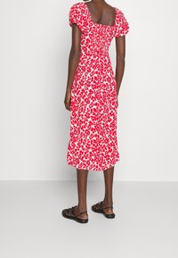 Rotes Blumenkleid mit Puffärmeln, quadratischem Rückenteil und gerafftem Taillenzug. Mittellang und aus leichtem Stoff, kombiniert mit schwarzen, flachen Sandalen.