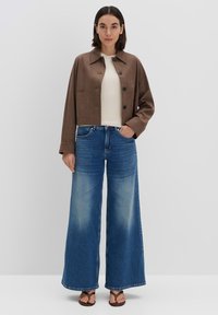 Braune kurze Jacke mit Knöpfen, cremefarbenes Oberteil und weit geschnittene blaue Jeans. Sandalen runden das Outfit ab. Schlichte, moderne Ästhetik.