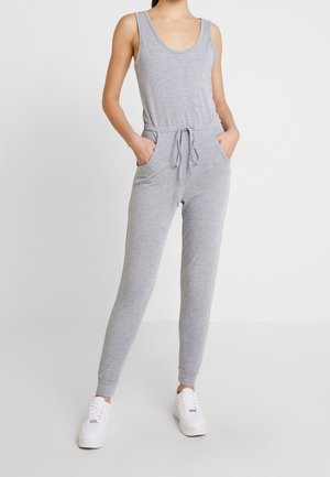 Femme portant une combinaison sans manches gris clair avec une taille à cordon et des baskets blanches, debout les mains dans les poches sur un fond blanc.