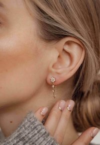 Boucles d'oreilles en forme de fleur en or avec une chaîne tombante ornée de petits gemmes clairs. Un pull en maille texturée est visible au premier plan.