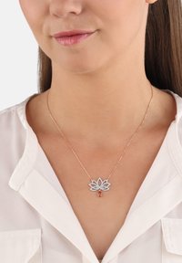 Frau trägt eine roségoldene Halskette mit einem lotusförmigen Diamanten-Anhänger und einem tropfenförmigen orangefarbenen Edelstein, kombiniert mit einer weißen Bluse.