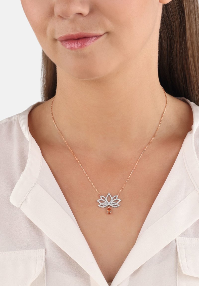 Frau trägt eine roségoldene Halskette mit einem lotusförmigen Diamanten-Anhänger und einem tropfenförmigen orangefarbenen Edelstein, kombiniert mit einer weißen Bluse.