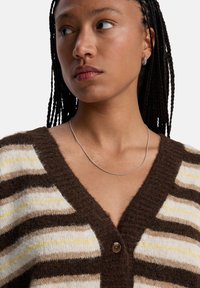 Jonge vrouw met gevlochten haar, gekleed in een bruin, crème en gele gestreepte cardigan en een zilveren ketting, die opzij kijkt.