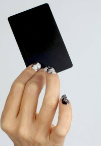 Carte de crédit noire tenue entre des doigts avec de longs ongles naturels. Les ongles présentent une base noire avec des motifs floraux blancs et des pointes effilées.