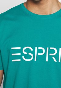 Tyrkysové bavlněné tričko s bílým potiskem textu "ESPRIT" na přední straně, kulatým výstřihem a jemnou texturou.