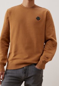 Hnědá mikina vyrobená z měkké tkaniny, s kulatým výstřihem, žebrovanými manžetami a černým logem ve tvaru diamantu na hrudi.