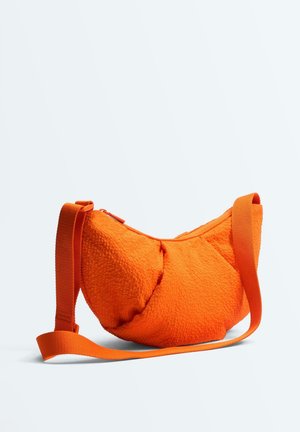 Jasne oranžová textúrou zdobená taška cez rameno s nastaviteľným popruhom a horným zapínaním na zips, zobrazená na jednoliatom svetlom pozadí.