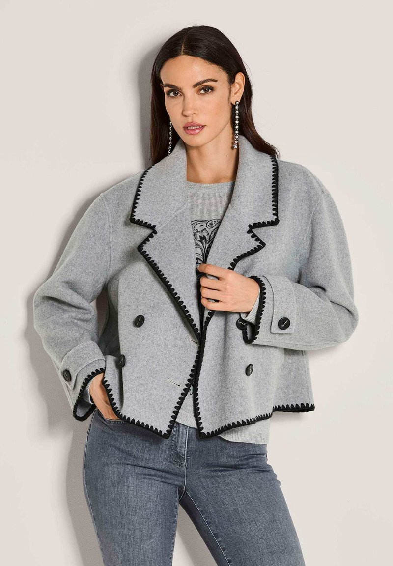 Vrouw in een grijze wol cropped jack met zwarte afwerking en knopen, gecombineerd met een grijs patroon top en strakke blauwe spijkerbroek, staande tegen een beige muur.