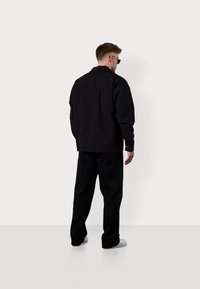 Black cotton jacket with a relaxed fit, featuring a collar and long sleeves. Paired with black pants and white sneakers. Rear view perspective.