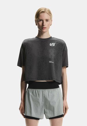 Femme portant un haut de course noir court avec les inscriptions « RUN » et « Swiss Engineering », assorti à un short de course superposé gris clair.