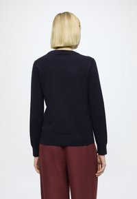 Tengerészkék kötött pulóver hosszú ujjakkal és kerek nyakkivágással, bordázott szegéllyel. Bordó, bő szárú nadrággal viselve.