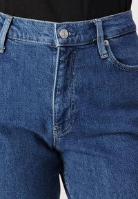 Jeans od plavog denima viskog struka s klasičnim dizajnom pet džepova, srebrnim dugmetom i kontrastnim šavovima duž šavova.
