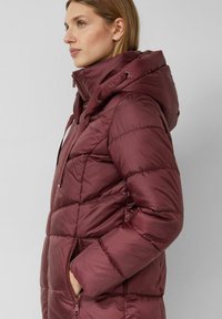 Donkerrode pufferjas met een doorgestikt ontwerp, vastgenaaide capuchon, ritssluiting aan de voorkant, zijzakken en trekkoorddetails aan de capuchon.
