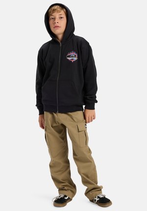 Hoodie noir zippé avec graphique de flammes et logo ; associé à un pantalon cargo beige avec poches latérales ; baskets noires avec lacets blancs.