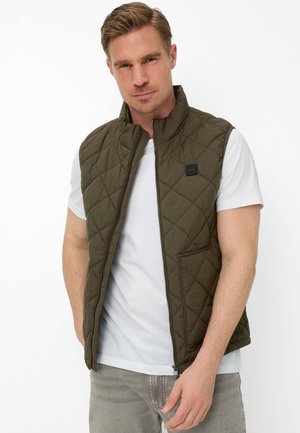 Man draagt een doorgestikte olijfgroene bodywarmer over een wit t-shirt en grijze spijkerbroek, staat met handen gedeeltelijk in de zakken tegen een witte achtergrond.