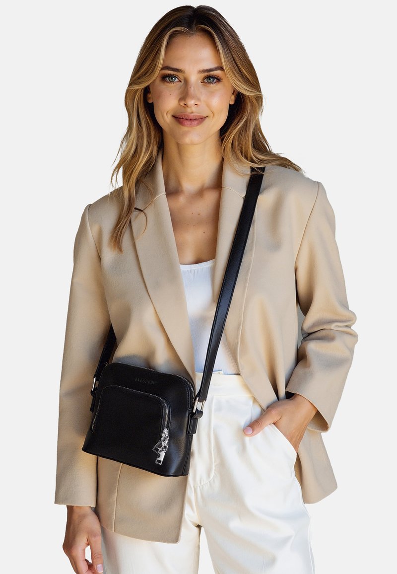 Borsa a tracolla in pelle nera con chiusura a zip, texture liscia, accessori in argento e tracolla regolabile. Le dimensioni suggeriscono un formato compatto.